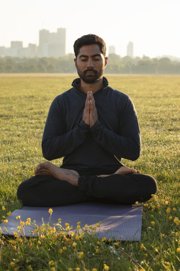 A man with a beard sits cross-legged in a meditative pose on a purple yoga mat in a grassy park in Kolkata, with a city skyline visible in the hazy background. The sunlight is golden, and there are small yellow flowers scattered around him